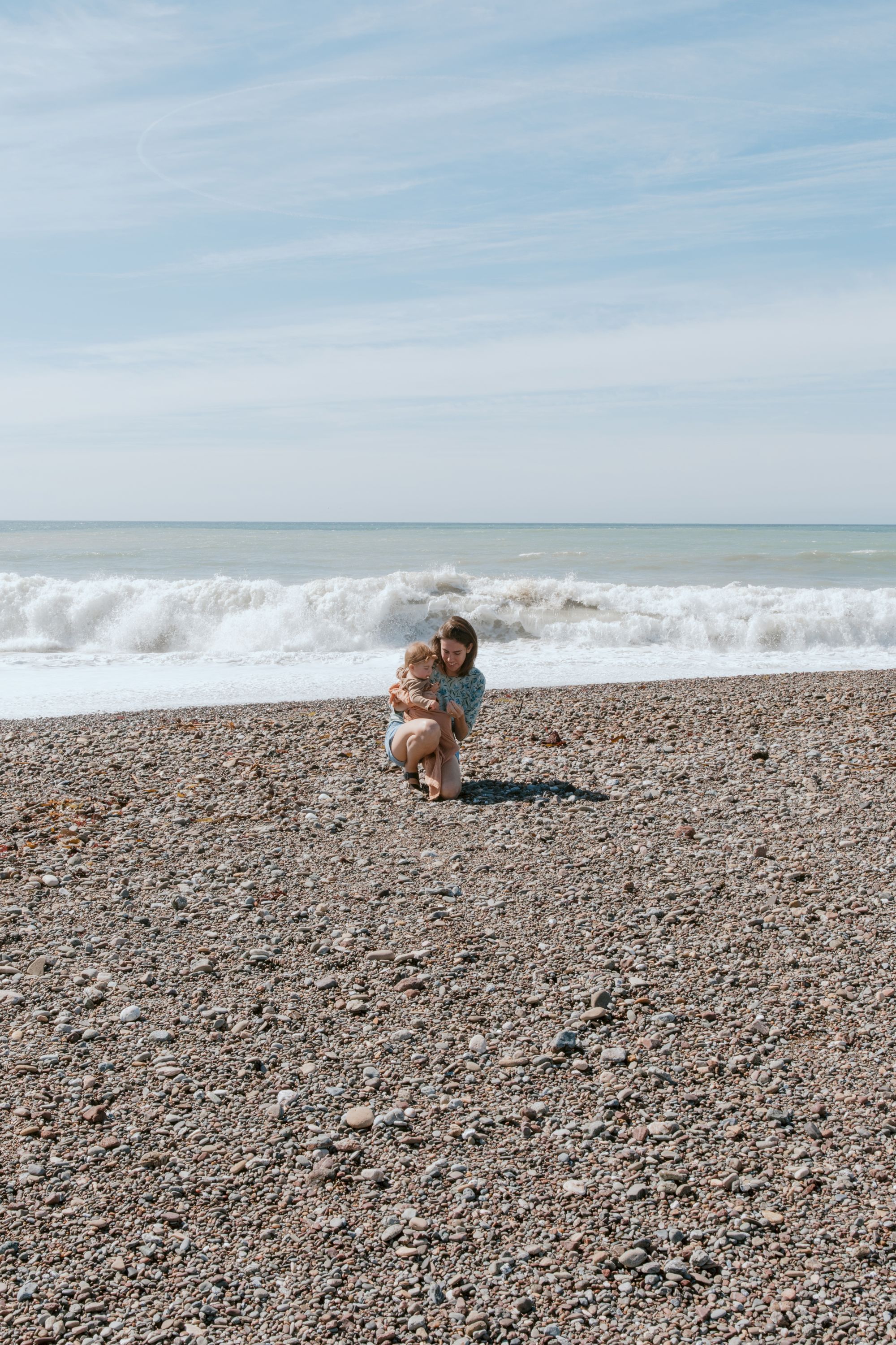 A person sits on a pebbly beach near gentle waves, hugging a small dog as the sea and sky meet at the horizon.
