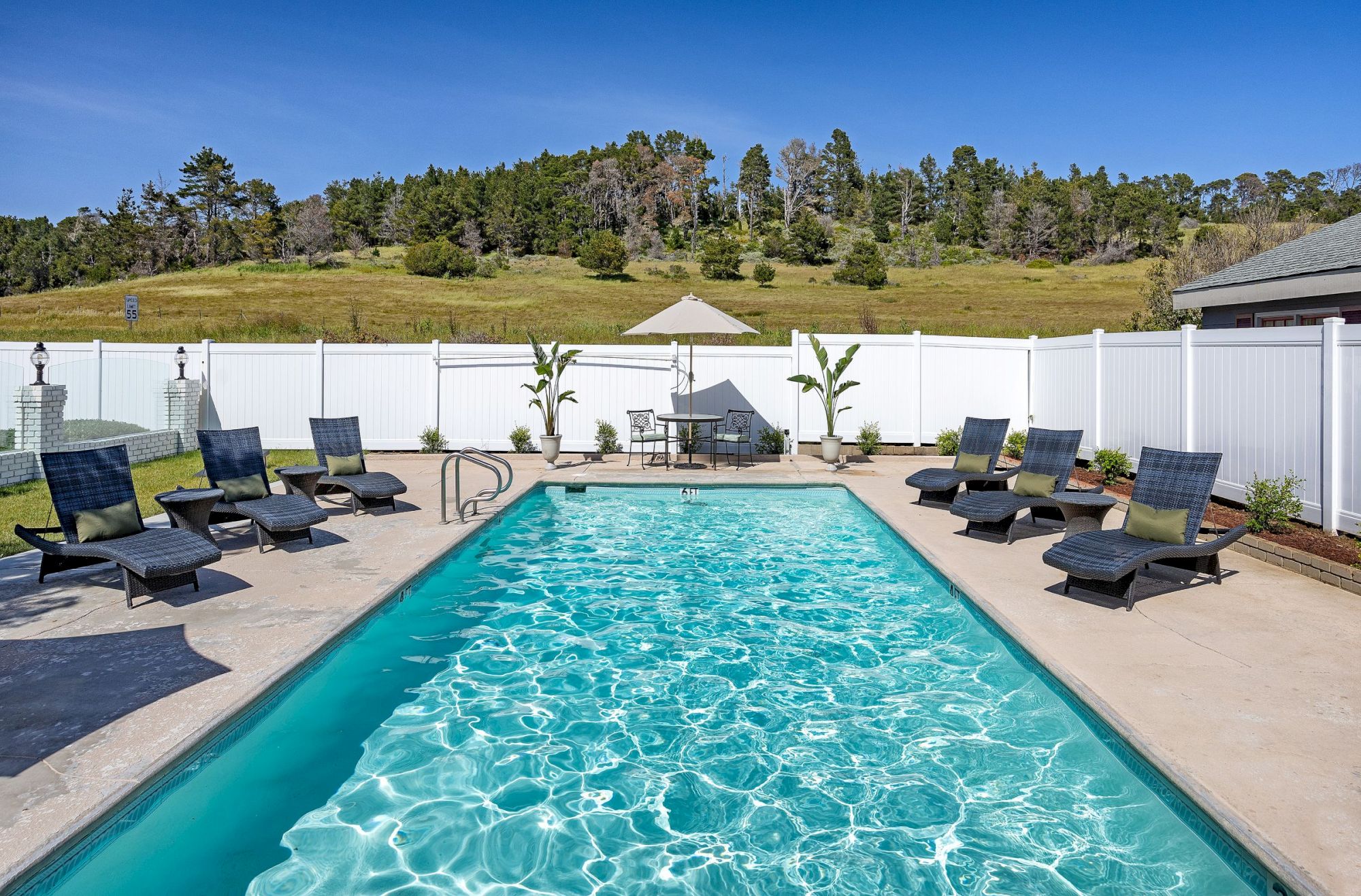 A sunny backyard pool surrounded by lounge chairs and umbrellas, with a white fence and a scenic hillside in the background.