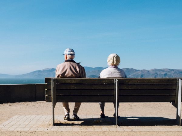 Two people sitting on a bench overlooking a vast landscape with mountains under a clear blue sky.