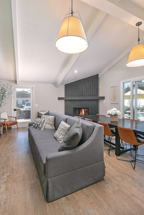 Modern living room with a gray sofa, built-in media cabinet, cozy fireplace, bright dining area, large windows, and a view of a pool/outdoor area.