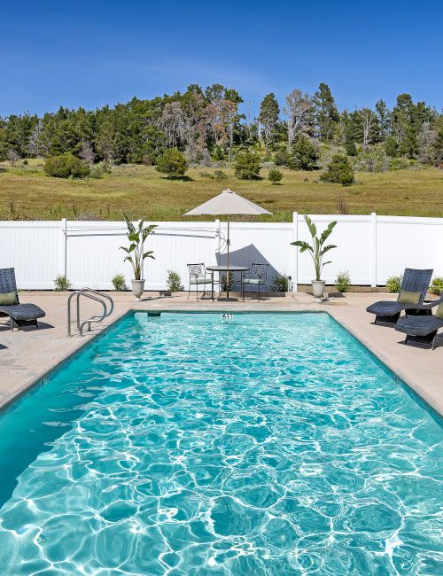 A rectangular backyard pool with clear blue water, surrounded by lounge chairs, a white fence, and a sunny, open sky. ending now.