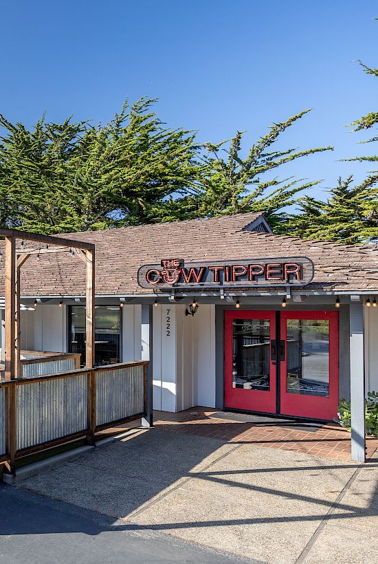A small restaurant with a wooden-frame outdoor patio, red front doors, patio tables, umbrellas, and shaded seating under a clear blue sky.