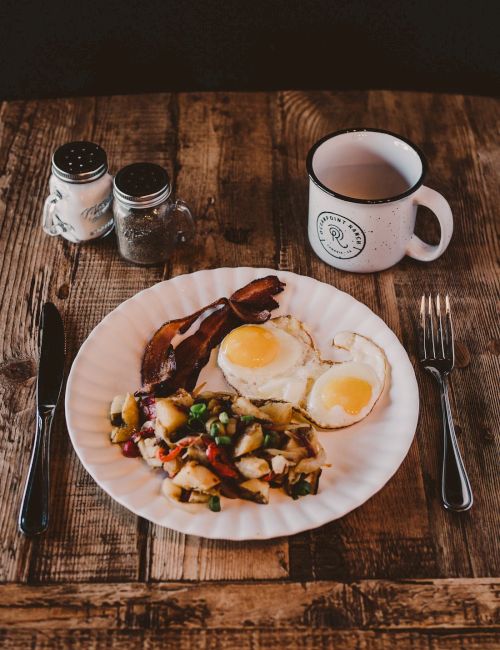 A hearty breakfast with sunny-side eggs and roasted veggies on a plate, beside a mug, salt and pepper shakers on a rustic wooden table.