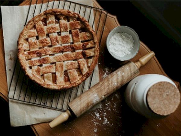 A lattice-topped pie on a cooling rack, surrounded by a rolling pin, a bowl with flour, and a jar, resting on a wooden surface.