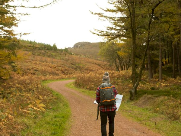 A person wearing a plaid jacket and backpack walks a winding dirt trail through a forest with autumn foliage, map in hand, heading away.
