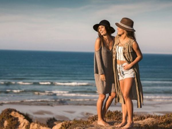 Two women stand on a sandy beach near the sea, wearing sun hats and stylish beach outfits, chatting and enjoying the coastal scenery.