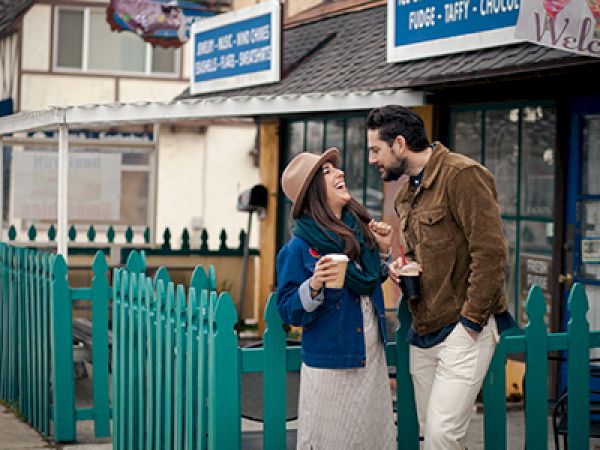 A couple is laughing together while holding coffee cups, standing near a teal fence outside shops with a warm and cozy ambiance.