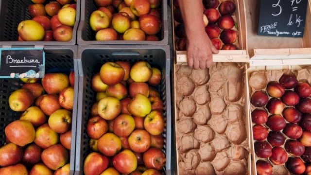 A market stall displays various apples in crates, with prices noted on chalkboards, and a person arranging them.