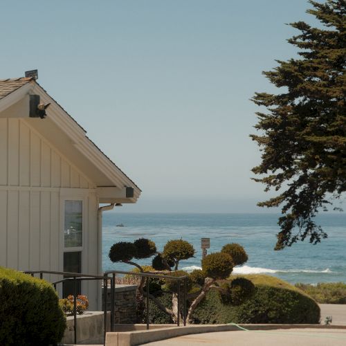 A white coastal house by a path, trimmed bushes, and view of the ocean with a large tree on the right.