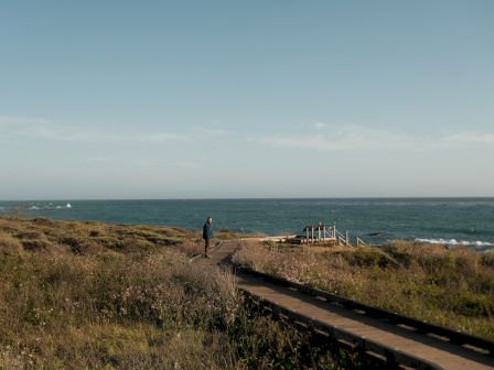 A person walks along a coastal path toward the sea, with grassy dunes, a wooden boardwalk, and a calm ocean under a clear sky.