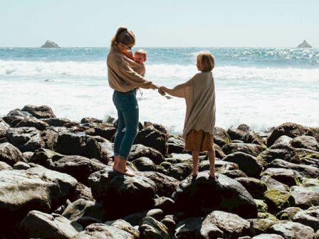 A person holding a baby and another child stand on rocks by the ocean, with waves breaking in the background.