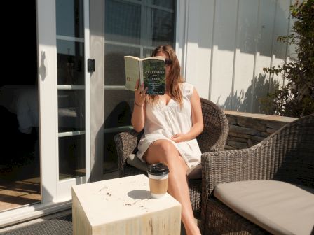A woman sits on an outdoor chair by a sliding door, reading a book with a coffee cup on a small wooden table beside her.