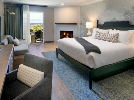 A cozy hotel room with a bed, fireplace, TV, desk, sofa, and open doors leading to a balcony with an ocean view.