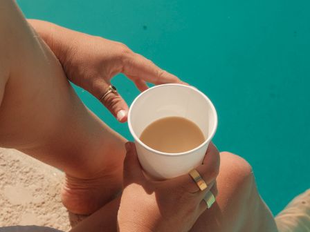 A person sits by a pool, holding a cup of coffee or tea, enjoying a sunny moment with legs dipped in bright blue water.