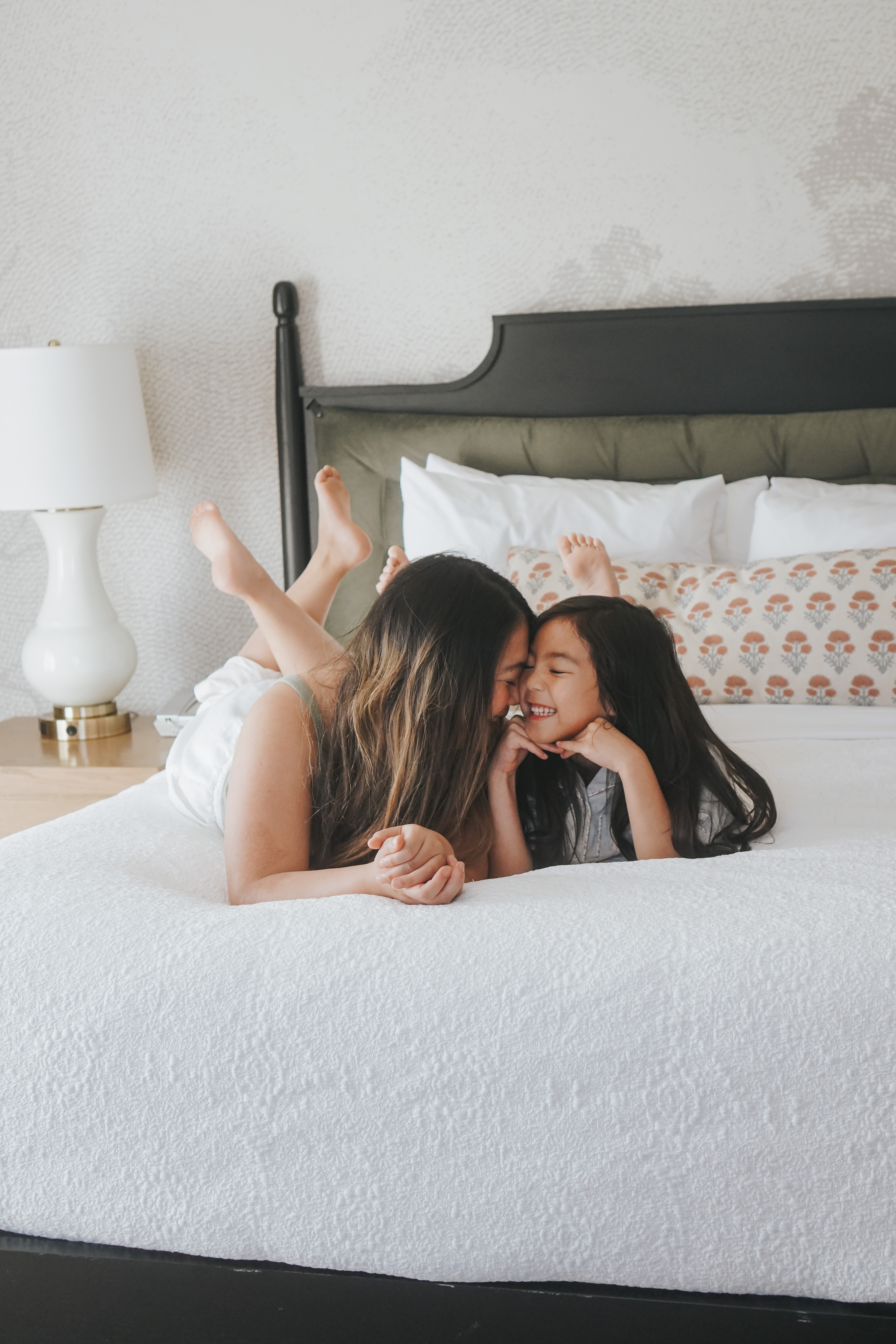 A woman and a young girl lying on a bed with white bedding and a black headboard.