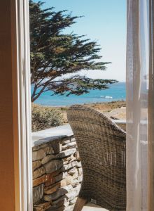 A seaside view through a doorway: a wicker chair on a stone patio, blue ocean beyond, sunny, with clear sky.