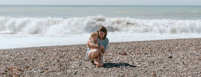 A child sits on a pebbly beach near the shore, with waves breaking behind them, under a cloudy sky.