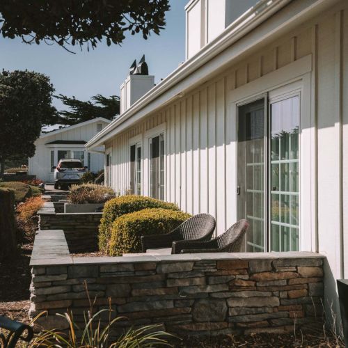 A small house with a white porch, green window frames, a short brick wall, and a tree casting shade, with some plants and a sunny sky.