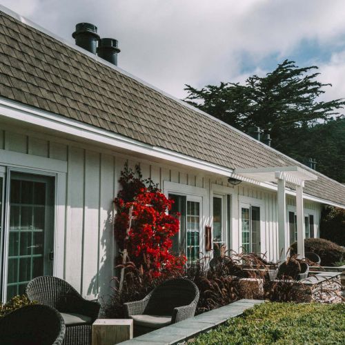 A single-story house with a stone porch, potted plants, and a red flowering shrub along the front, beneath a cloudy blue sky.