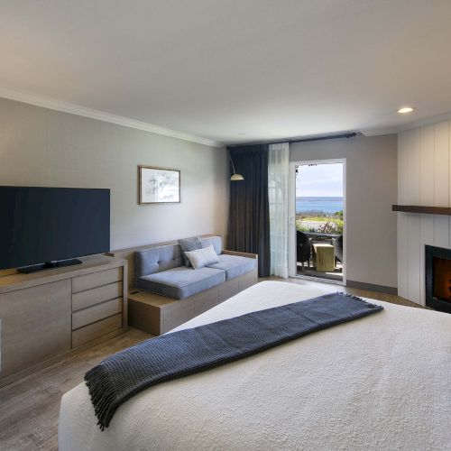 A bright hotel room with a large bed, sofa, TV on a dresser, fireplace, and a balcony view of the outdoors. Ending with a period.