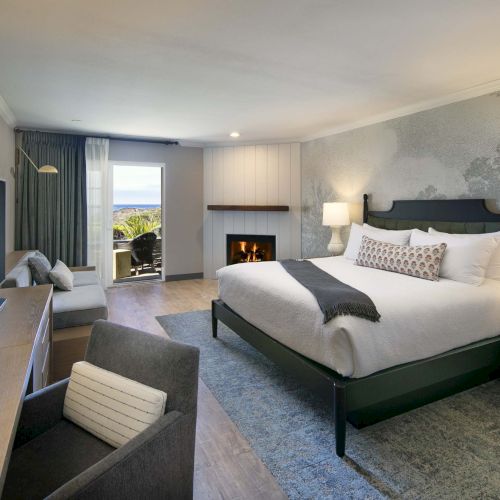 A spacious hotel room with a king bed, two nightstands, a desk and chair, a sitting area, a fireplace, and a balcony overlooking scenery.