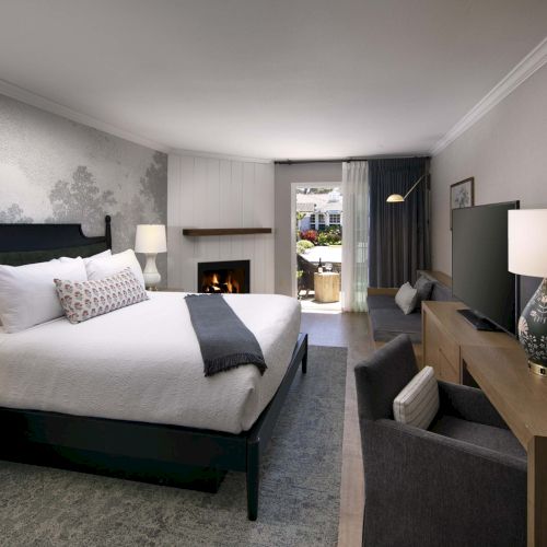 A modern hotel room with a large bed, nightstands, a desk area, a fireplace, seating, and soft lighting&mdash;cozy and well-appointed.