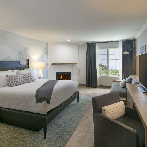 A hotel room with a large bed, nightstands, lamps, a fireplace, a desk and chair, a sofa, and a window with dark curtains.