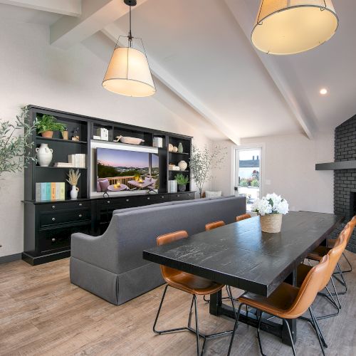 A modern, open-concept living and dining room with a gray sofa, black cabinets, wood dining table, pendant lights, and a fireplace.