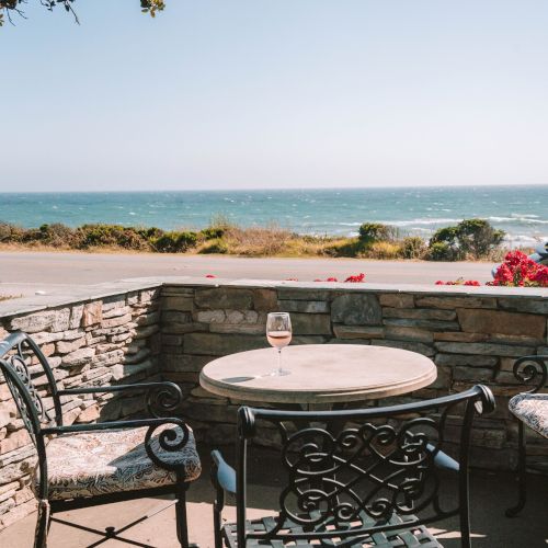 A seaside patio with a wrought-iron table and chairs, a glass of wine, stone railing, and a bright ocean view under a clear sky, peaceful.
