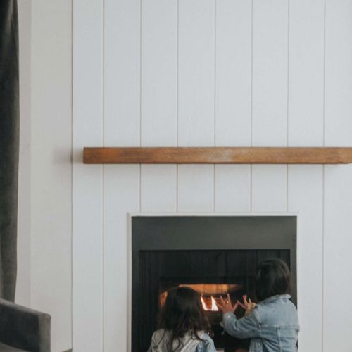 A cozy living space with a white paneled wall, a wooden mantel around a black fireplace, and a small group of people sitting on the floor by the hearth.