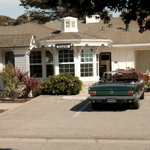 A beige suburban house with a dark car parked in the driveway, a well-kept lawn, and a tree partially framing the scene.