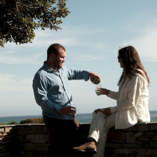 Two people sit on a railing by the beach, chatting as the sun sets behind them.