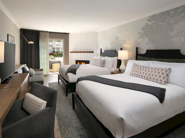 A stylish hotel room with two plush queen beds, a desk and chairs, a seating area, and a balcony door letting in soft light. End sentence.