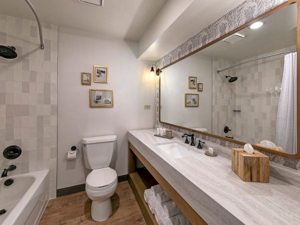 A modern bathroom with a bathtub/shower combo, white fixtures, a long marble countertop, a large framed mirror, and simple decor, including a tissue box and framed art.