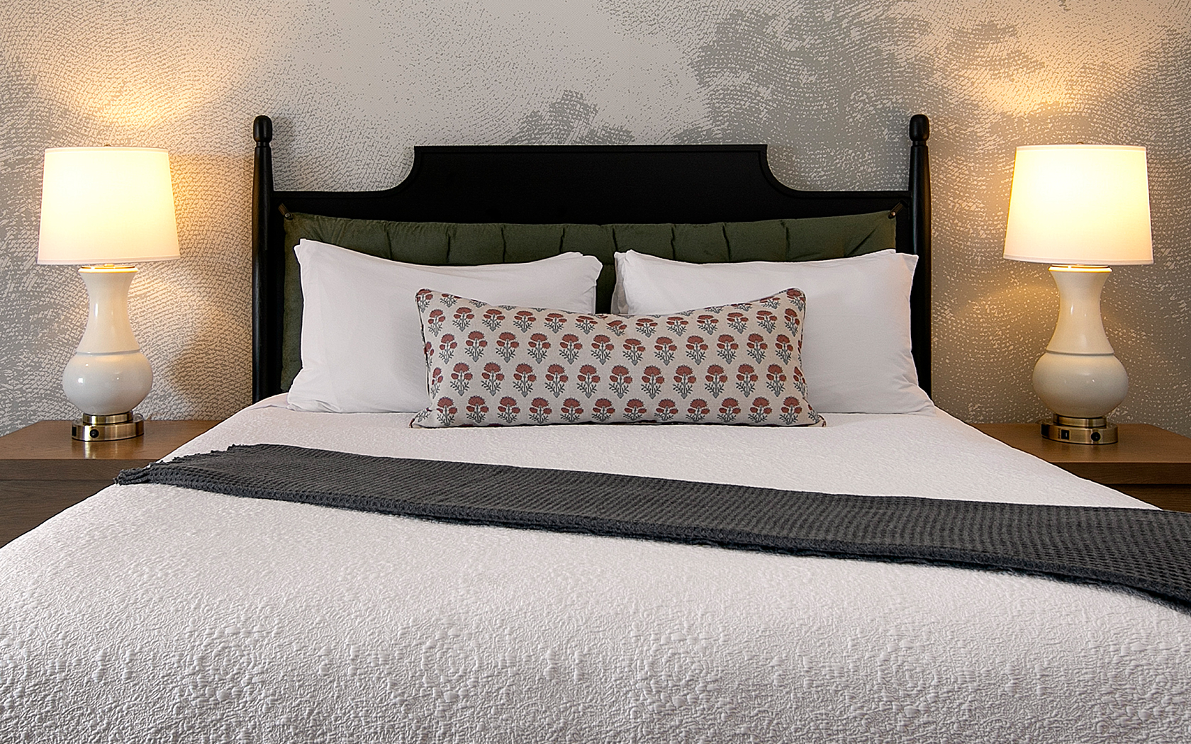 A neatly made bed with white bedding, two wooden nightstands with lamps, and patterned pillows.