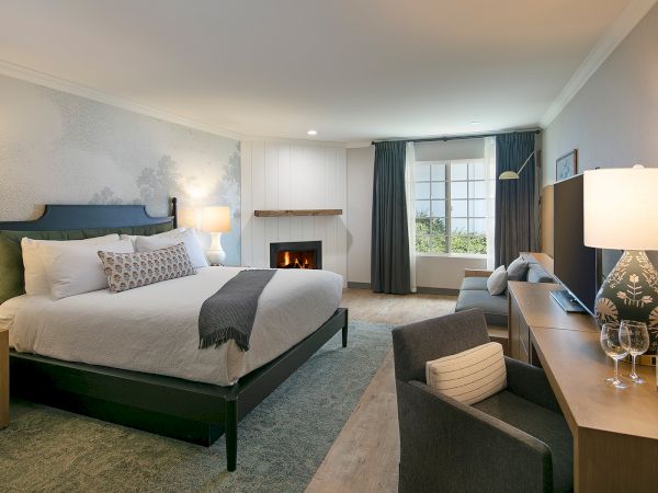 A spacious hotel room with a king bed, blue-gray decor, desk, seating area, fireplace, and large window with dark curtains.