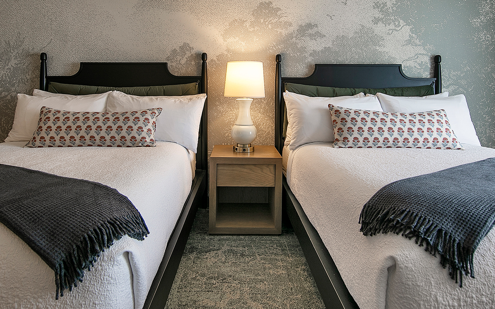 A hotel room with two beds, a bedside table, and a lamp.