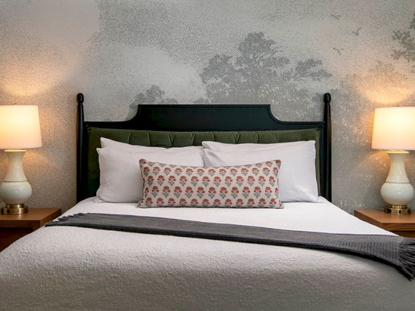 A neatly made bed with white linens, two nightstands, matching lamps, a decorative bolster pillow, and a soft gray blanket at the foot.