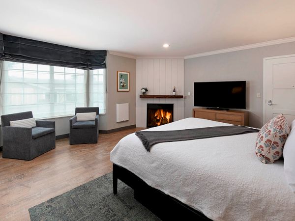 A modern hotel-style bedroom with a fireplace, large window seating, a flat-screen TV, and a neatly made bed with decorative pillows.