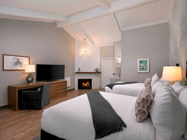 A modern hotel room with two white beds, a fireplace, a TV, a wooden dresser, and soft lighting, creating a cozy, contemporary vibe.