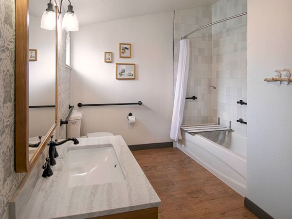 A bathroom with a double sink vanity, large mirror, wood-framed mirror, white countertops, and a bathtub/shower combo with a glass door, white tile, and beige walls.